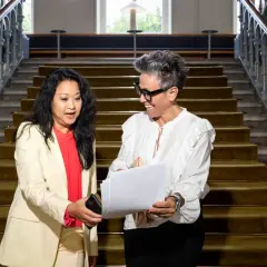 Två kvinnor diskuterar papper vid en trappa. Den ena kvinnan, klädd i ljus kavaj, ger en telefon till den andra, som ler och håller i dokumenten. Kanske en personlig assistent som hjälper till med organisationen.