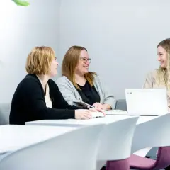 Tre kvinnor diskuterar sitt gymnasiearbete på Komvux vid ett bord med böcker och en laptop.