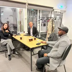 Tre personer sitter vid ett bord och diskuterar juridik nyheter. En kvinna gestikulerar, en man i kostym lyssnar uppmärksamt och en man med keps ser fundersam ut.
