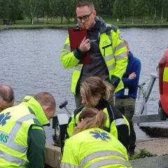 Ambulanssjukvårdare i utbildning distans övar vid vattnet. De bär reflexvästar och en instruktör antecknar.