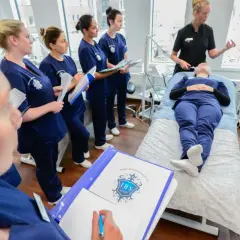 Elever på hudterapeut utbildning distans lyssnar på instruktör. En elev ligger på en brits medan andra antecknar.