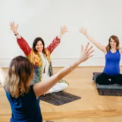 Tre kvinnor sitter på yogamattor med armarna uppåt. En av dem är gravid. En inspirerande scen från en yoga utbildning.