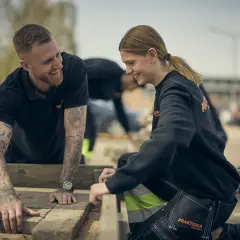 Två glada ungdomar, klädda i arbetskläder från Praktiska Gymnasiet, lägger stenplattor utomhus. En bra start på ett jobb efter gymnasiet!