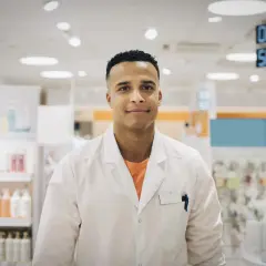 Ung man i vit rock, redo för apotekstekniker utbildning. Bakom honom syns hyllor med produkter i ett apotek.