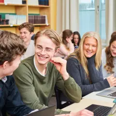 Glada elever på ett idrottsgymnasium i Stockholm skrattar tillsammans vid sina datorer i ett klassrum.