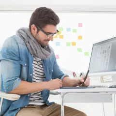 En grafisk designer arbetar vid sitt skrivbord med en ritplatta och dator. Färgade post-it-lappar sitter på väggen bakom.