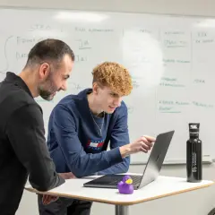 Två män diskuterar kod på en laptop. En .net utvecklare pekar på skärmen medan en kollega tittar på. En whiteboard med anteckningar syns i bakgrunden.