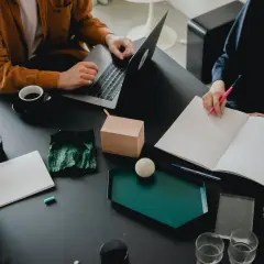 Två personer vid ett bord, en med laptop, den andra skriver i en bok. Kaffe och anteckningsmaterial. De är bi utvecklare som samarbetar.