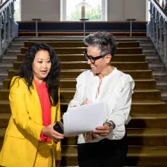 Två kvinnor diskuterar papper i en trappa. Den ena, klädd i gult, verkar vara en kreativ chef som ger instruktioner.