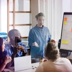 Mötesdeltagare diskuterar en presentation om hållbarhet, liknande en byggnadsantikvarie utbildning. En kvinna leder diskussionen vid en whiteboard.