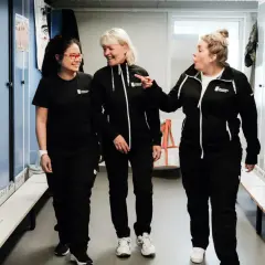 Tre kvinnor i svarta träningskläder skrattar i ett omklädningsrum. De verkar vara på en lokalvårdare utbildning.