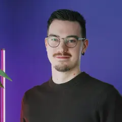 En ung man med glasögon och mustasch ler mot kameran. Bakgrunden är lila med en röd vägg och en växt. Han kanske är student på Chas Academy.