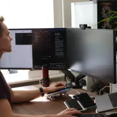 Man vid datorn, fokuserad på sin programmerings kurs. Flera skärmar visar kod och text. En mikrofon och en webbkamera är synliga.