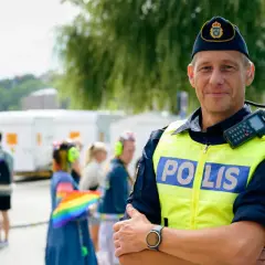 Polis i tjänst, kanske på väg att plugga till polis. Han bär gul reflexväst med texten "POLIS" och en keps med Sveriges riksvapen. I bakgrunden syns folk och en regnbågsflagga.