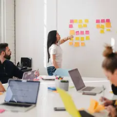 Teamwork i ett ljust kontor. En kvinna pekar på post-it-lappar på en vägg, medan andra diskuterar. Kanske en embedded utvecklare som presenterar idéer.