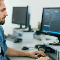 Manlig IT-tekniker med uppsatt hår och skägg arbetar vid två datorskärmar med kod.