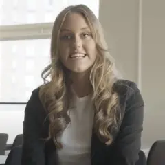 En ung kvinna med blont, lockigt hår ler och talar. Hon bär en svart kavaj över en vit topp. Bakom henne syns fönster och stolar, kanske i en lokal på Katedralskolan, Uppsala.
