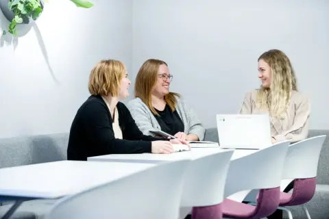 Tre kvinnor diskuterar sitt gymnasiearbete på Komvux vid ett bord med böcker och en laptop.