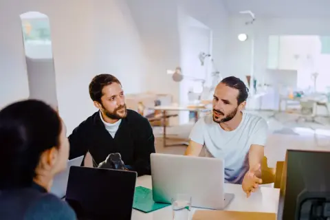 Män och kvinnor diskuterar webbutveckling utbildning vid laptops i ett ljust kontorslandskap.