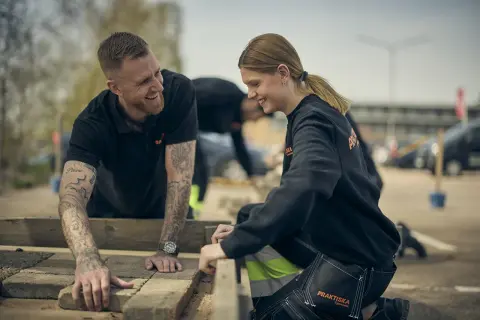 Två glada ungdomar, klädda i arbetskläder från Praktiska Gymnasiet, lägger stenplattor utomhus. En bra start på ett jobb efter gymnasiet!