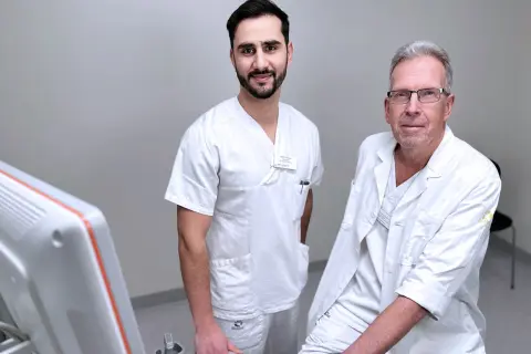 Två män i vita arbetskläder står bredvid en ultraljudsmaskin. Den yngre mannen ler mot kameran, den äldre har glasögon. De är en del av sonograf utbildning.