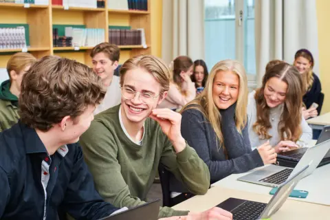 Glada elever på ett idrottsgymnasium i Stockholm skrattar tillsammans vid sina datorer i ett klassrum.