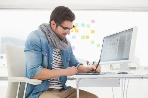 En grafisk designer arbetar vid sitt skrivbord med en ritplatta och dator. Färgade post-it-lappar sitter på väggen bakom.