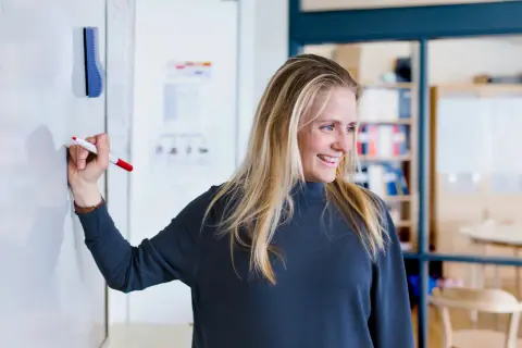 Glad kvinna med blondt hår skriver på en whiteboard. Hon kanske studerar socialpedagog utbildning distans.