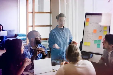 Mötesdeltagare diskuterar en presentation om hållbarhet, liknande en byggnadsantikvarie utbildning. En kvinna leder diskussionen vid en whiteboard.