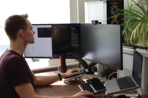 Man vid datorn, fokuserad på sin programmerings kurs. Flera skärmar visar kod och text. En mikrofon och en webbkamera är synliga.