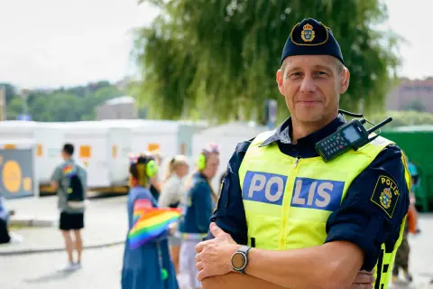 Polis i tjänst, kanske på väg att plugga till polis. Han bär gul reflexväst med texten "POLIS" och en keps med Sveriges riksvapen. I bakgrunden syns folk och en regnbågsflagga.