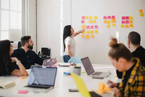 Teamwork i ett ljust kontor. En kvinna pekar på post-it-lappar på en vägg, medan andra diskuterar. Kanske en embedded utvecklare som presenterar idéer.