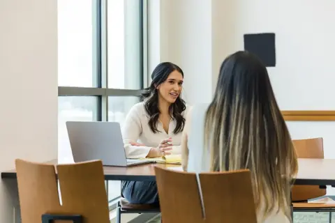 Två kvinnor sitter vid ett bord, en med laptop. Kanske diskuterar de kurator utbildning distans.