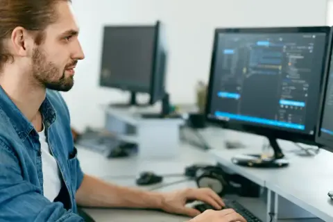 Manlig IT-tekniker med uppsatt hår och skägg arbetar vid två datorskärmar med kod.
