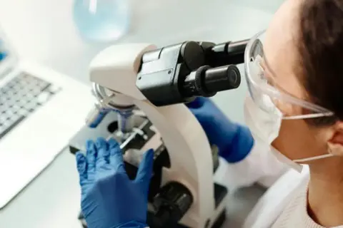 En forensiker arbetar med ett mikroskop i ett laboratorium. En laptop syns i bakgrunden.
