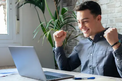 Ung man jublar framför sin laptop, kanske har han hittat roliga jobb eller lyckats med ett projekt.