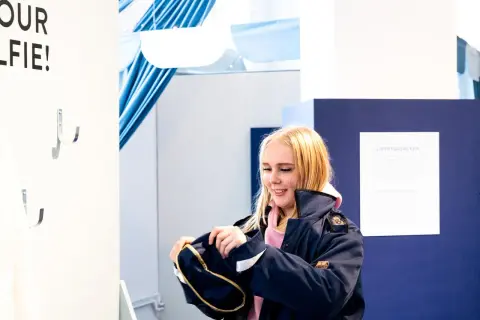 Glad tjej med ljusblont hår, klädd i mörkblå jacka, tittar ner på en väska. Hon är på ett event för ett vf gymnasieprogram.