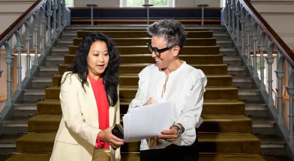 Två kvinnor diskuterar papper vid en trappa. Den ena kvinnan, klädd i ljus kavaj, ger en telefon till den andra, som ler och håller i dokumenten. Kanske en personlig assistent som hjälper till med organisationen.