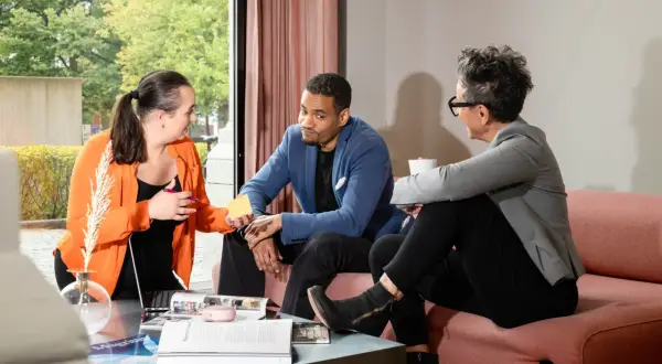 Tre personer diskuterar, en kvinna i orange ger en gul post-it till en man i blå kavaj. Kanske planerar de en virtuell assistent utbildning.