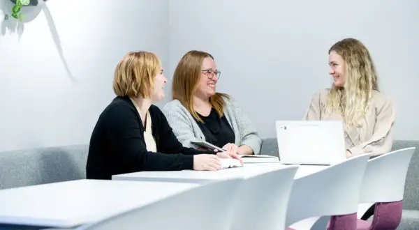 Tre kvinnor diskuterar sitt gymnasiearbete på Komvux vid ett bord med böcker och en laptop.
