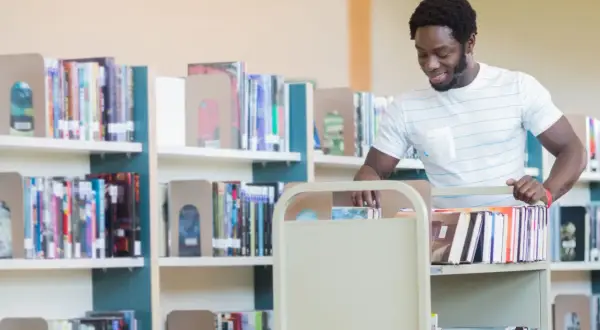 Man med bokvagn i bibliotek, kanske på väg att sortera böcker som en del av sin bibliotekarie utbildning.