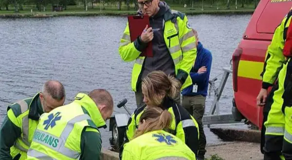 Ambulanssjukvårdare i utbildning distans övar vid vattnet. De bär reflexvästar och en instruktör antecknar.