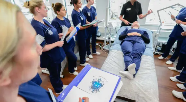 Elever på hudterapeut utbildning distans lyssnar på instruktör. En elev ligger på en brits medan andra antecknar.
