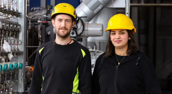 Två personer med gula hjälmar står framför industriell utrustning. De är studenter vid byggbranschens utbildningscenter.