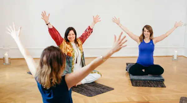 Tre kvinnor sitter på yogamattor med armarna uppåt. En av dem är gravid. En inspirerande scen från en yoga utbildning.