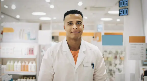 Ung man i vit rock, redo för apotekstekniker utbildning. Bakom honom syns hyllor med produkter i ett apotek.