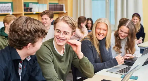 Glada elever på ett idrottsgymnasium i Stockholm skrattar tillsammans vid sina datorer i ett klassrum.