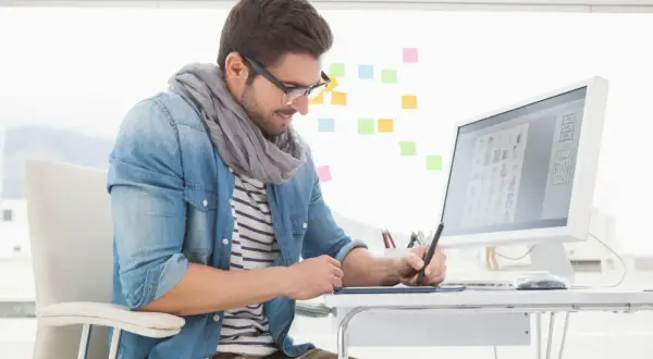 En grafisk designer arbetar vid sitt skrivbord med en ritplatta och dator. Färgade post-it-lappar sitter på väggen bakom.