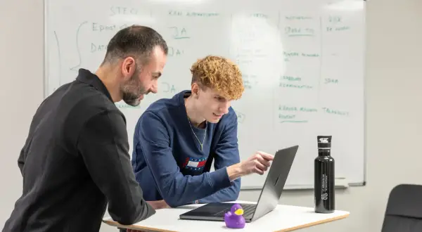 Två män diskuterar kod på en laptop. En .net utvecklare pekar på skärmen medan en kollega tittar på. En whiteboard med anteckningar syns i bakgrunden.
