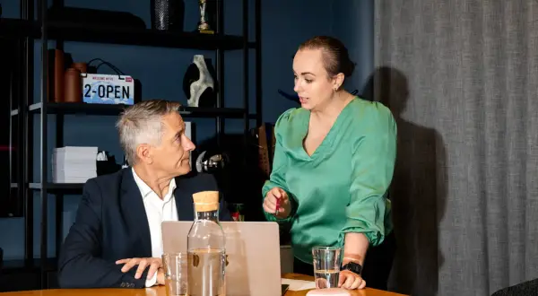 En kvinna i grön blus diskuterar med en man i kostym vid ett bord. De verkar samarbeta som payroll business partners.